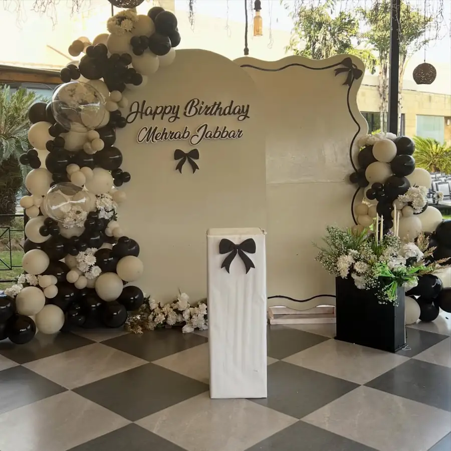 Black, white and gold balloon arch around a cream backdrop on a chequered floor