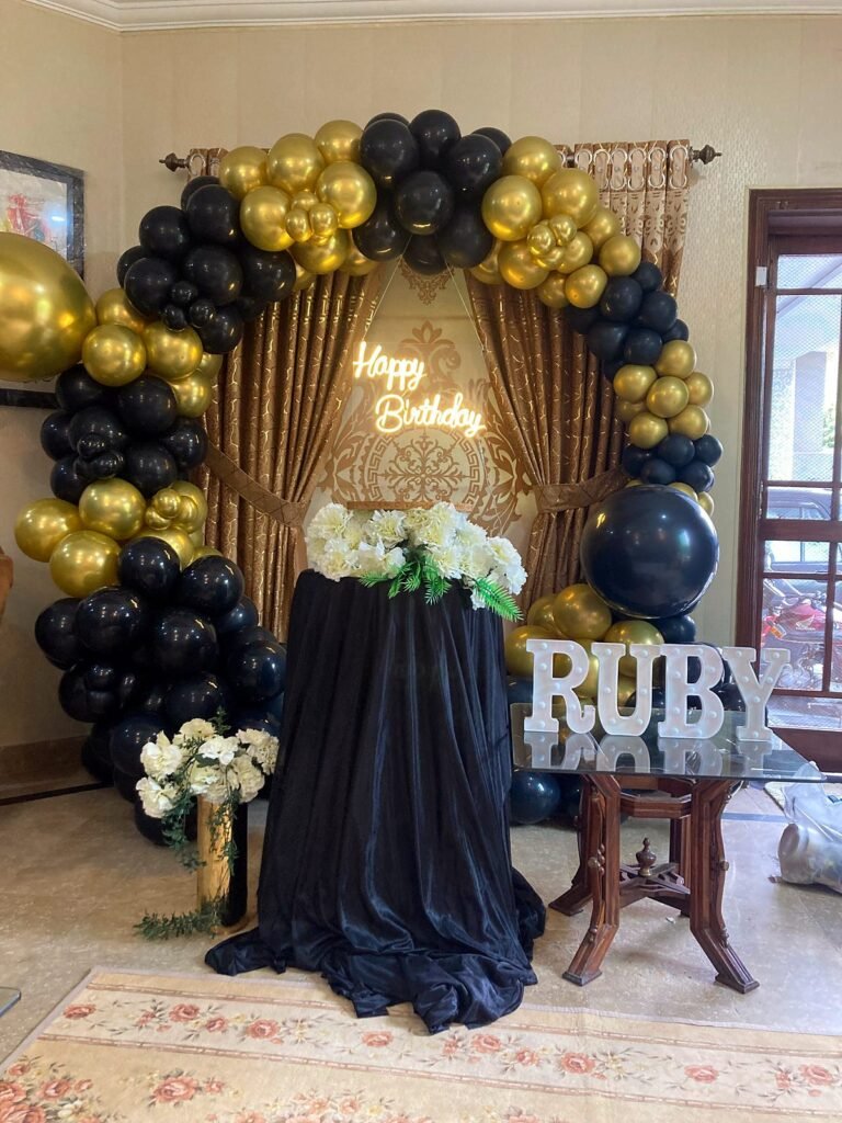 Black and gold balloon ring with a Happy Birthday neon sign and name letters on a table