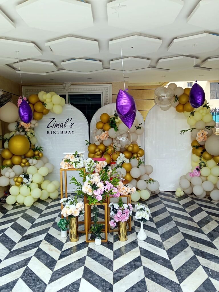 Gold and white birthday arch with flower stands and purple balloons